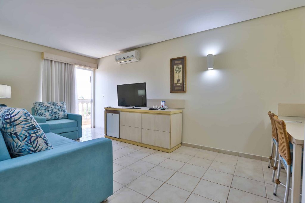 Suíte Master – Sala Espaçosa sala de estar da Suíte Master no Thermas de Olímpia Resorts by Mercure. Decoração elegante com sofá e poltrona em tons acolhedores. Frigobar para uso livre e TV a cabo disponível para os hóspedes. Ambiente naturalmente refrigerado e com ar-condicionado. Móvel de organização e mesa para comodidade dos visitantes. O refúgio perfeito para relaxar e desfrutar de momentos memoráveis durante a estadia.