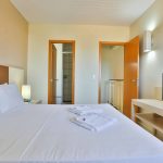 Piso Superior: Suíte principal com cama de casal e decoração elegante, iluminada por uma janela. Conte também com um banheiro exclusivo da Suíte.