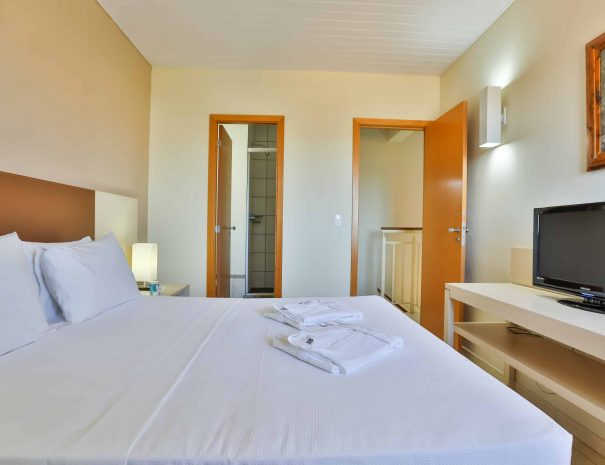 Piso Superior: Suíte principal com cama de casal e decoração elegante, iluminada por uma janela. Conte também com um banheiro exclusivo da Suíte.